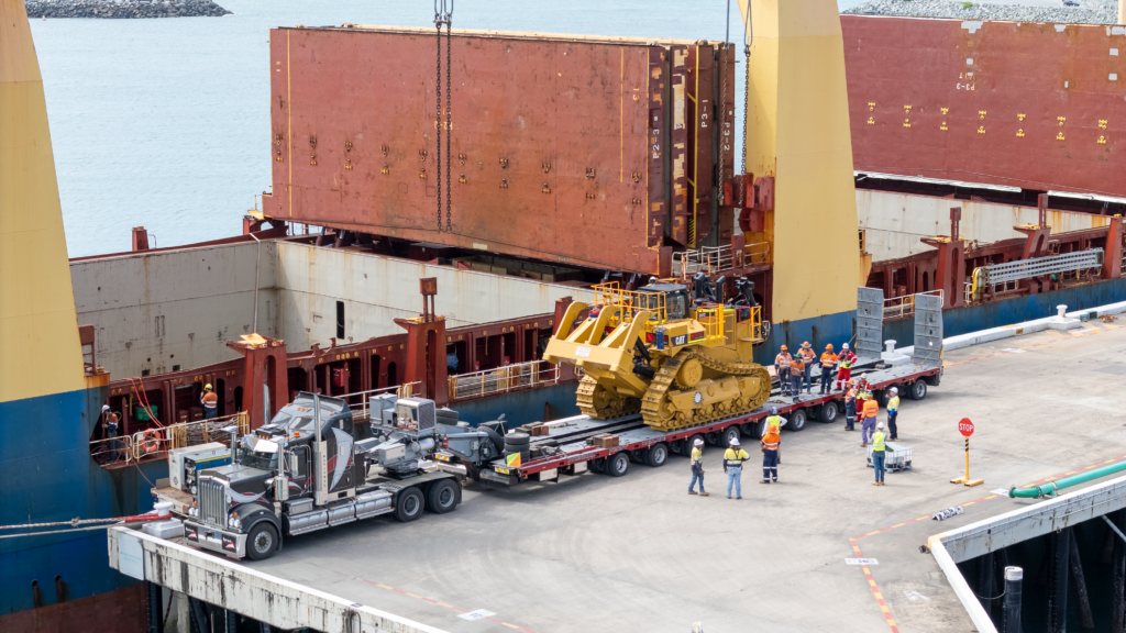 A Caterpillar D11 dozer sits secured on a National Heavy Haulage low-loader truck at a port, as workers prepare to load the oversized mining machine onto a cargo ship using heavy lifting equipment.