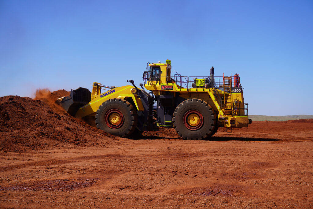 A Komatsu WA1200 wheel loader moves a large pile of red earth across an open mining site under clear blue skies, with dust rising from the bucket as it works.