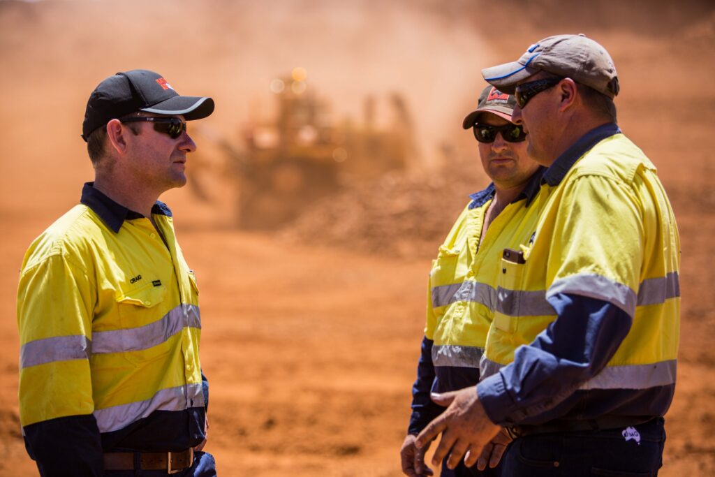 Three workers in high-visibility uniforms stand together discussing operations on an iron ore mining site, with heavy machinery blurred in the background.