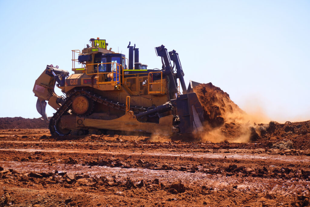 A Caterpillar D11T dozer pushes a heavy load of red earth across a dusty mining worksite under clear blue skies, with dirt and dust flying from the blade as the machine operates.